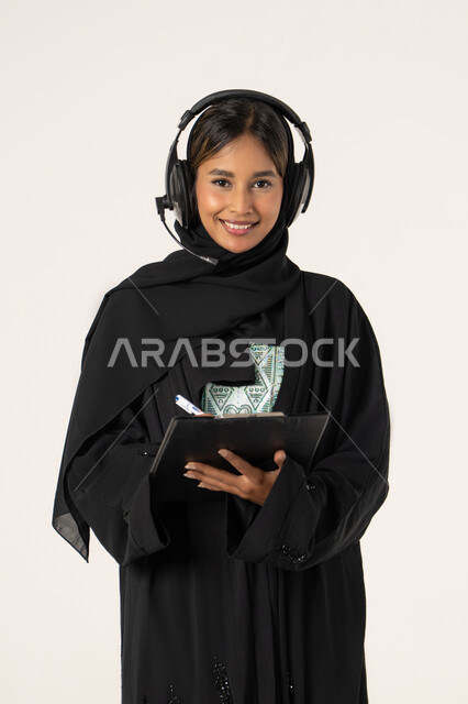 متابعة وتطوير العمل، مفهوم خدمة العملاء، تدوين و تسجيل الملاحظات و الشكاوي، بورتريه مقرب لموظفة عربية خليجية اماراتية محجبة مبتسمة ترتدي عباءة سوداء تضع سماعات الهيدفون على رأسها و تقوم بالكتابة على الاوراق، الرد على الاستفسارت والتساؤولات، خلفية بيضاء