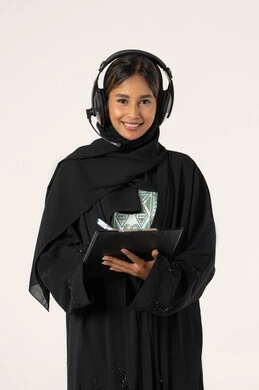 Follow-up and development of work, customer service concept, recording and registering notes and complaints, close-up portrait of a smiling veiled Emirati Gulf Arab female employee wearing a black abaya, putting headphones on her head and writing on papers, responding to inquiries and questions, white background