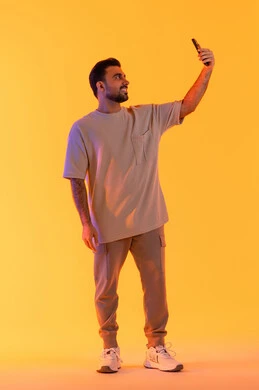 تقنيات معاصرة و تفاعل مع التكنولوجيا، التقاط الصور التذكارية وتوثيق اللحظات السعيدة, بورتريه لرجل عربي خليجي اماراتي يرتدي الزي الكاجوال يمسك الهاتف المحمول ويقوم بألتقاط صورة سيلفي، مفهوم الأناقة والرجولة والاهتمام بالمظهر الخارجي, بالطول الكامل، خلفية برتقاليه