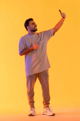 Interaction with technology and contemporary techniques, capturing memorable photos and documenting happy moments, a portrait of an Arab Gulf Emirati man wearing casual attire holding a mobile phone and taking a selfie, the concept of elegance, masculinity, and attention to appearance, full-length, with an orange background.