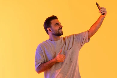 Capturing memorable photos and documenting happy moments, a portrait of an Arab Gulf Emirati man wearing casual attire holding a mobile phone and taking a selfie, the concept of elegance, masculinity, and attention to appearance, contemporary techniques and interaction with technology, orange background.