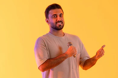 العروض الفنية المسرحية والفقرات الترفيهية، غناء مقطع صوتي غنائي، بورتريه لرجل عربي خليجي إماراتي يرتدي الزي الكاجوال يمسك المايكروفون ويغني بحماس، إيماءات الإندماج والتفاعل، إجراء المقابلات وطرح الأسئلة، التواصل الفعال وتبادل الحوارات، خلفية برتقالي .