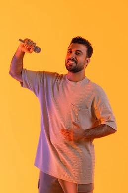 Singing a musical audio clip, a portrait of an Emirati Gulf Arab man wearing casual attire holding a microphone and singing enthusiastically, gestures of engagement and interaction, presenting artistic theatrical performances and entertainment segments, conducting interviews and asking questions, effective communication and exchanging dialogues, orange background.