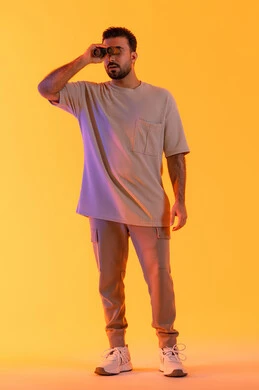 توضيح الرؤية وتقريبها، تعابير الاندماج والتركيز, رؤية الاشياء البعيدة عبر التلسكوب,  حب الاستكشاف و المغامرة, بورتريه لشاب عربي خليجي اماراتي مبتسم يرتدي الزي الكاجوال يحمل بيده منظار ثنائي العينين ينظر إلى شيئ ما، صورة الجسم بالطول الكامل, خلفية برتقاليه