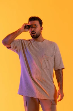 تقريب الرؤية وتوضيحها، تعابير الاندماج والتركيز, رؤية الاشياء البعيدة عبر التلسكوب,  حب الاستكشاف و المغامرة, بورتريه لشاب عربي خليجي اماراتي مبتسم يرتدي الزي الكاجوال يحمل بيده منظار ثنائي العينين ينظر إلى شيئ ما، التخطيط ووضع الأهداف المستقبلية، خلفية برتقاليه