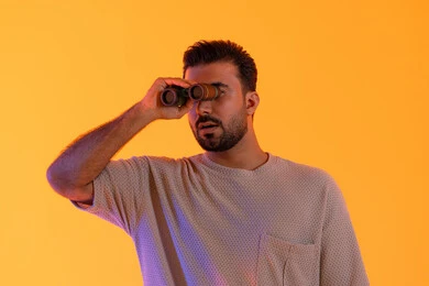 التخطيط ووضع الأهداف المستقبلية، تقريب الرؤية وتوضيحها، تعابير الاندماج والتركيز, رؤية الاشياء البعيدة عبر التلسكوب,  حب الاستكشاف و المغامرة, بورتريه لشاب عربي خليجي اماراتي مبتسم يرتدي الزي الكاجوال يحمل بيده منظار ثنائي العينين ينظر إلى شيئ ما، خلفية برتقاليه