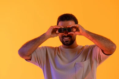 مفهوم حب الاستكشاف و المغامرة,  التخطيط ووضع الأهداف المستقبلية، تقريب الرؤية وتوضيحها، تعابير السعادة والسرور, رؤية الاشياء البعيدة عبر التلسكوب, بورتريه لشاب عربي خليجي اماراتي مبتسم يرتدي الزي الكاجوال يحمل بيده منظار ثنائي العينين ينظر إلى شيئ ما، خلفية برتقاليه