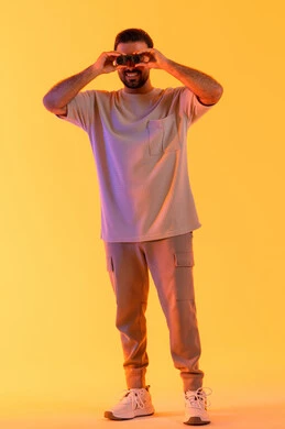 Gestures of enjoyment and pleasure, seeing distant things through a telescope, love for exploration and adventure, a portrait of a smiling young Arab Gulf Emirati man wearing casual attire holding binoculars in his hand looking at something, clarifying and zooming in on the view, full-body image, orange background.