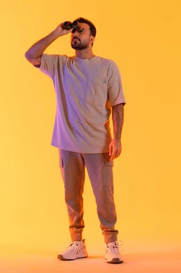 تعابير الاندماج والتركيز, رؤية الاشياء البعيدة عبر التلسكوب,  حب الاستكشاف و المغامرة, بورتريه لشاب عربي خليجي اماراتي مبتسم يرتدي الزي الكاجوال يحمل بيده منظار ثنائي العينين ينظر إلى شيئ ما، توضيح الرؤية وتقريبها، صورة الجسم بالطول الكامل, خلفية برتقاليه