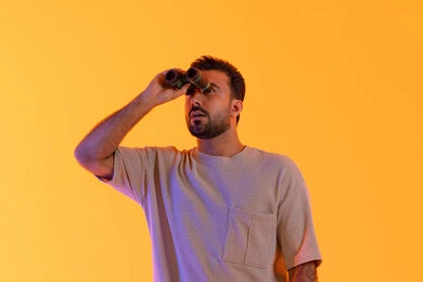 حب الاستكشاف و المغامرة,  التخطيط ووضع الأهداف المستقبلية، تقريب الرؤية وتوضيحها، تعابير الاندماج والتركيز, رؤية الاشياء البعيدة عبر التلسكوب, بورتريه لشاب عربي خليجي اماراتي مبتسم يرتدي الزي الكاجوال يحمل بيده منظار ثنائي العينين ينظر إلى شيئ ما، خلفية برتقاليه