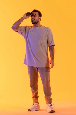 رؤية الاشياء البعيدة عبر التلسكوب, إيماءات الاستمتاع و السرور, حب الاستكشاف و المغامرة, بورتريه لشاب عربي خليجي اماراتي مبتسم يرتدي الزي الكاجوال يحمل بيده منظار ثنائي العينين ينظر إلى شيئ ما، توضيح الرؤية وتقريبها، صورة الجسم بالطول الكامل, خلفية برتقاليه