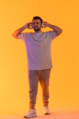 Standing confidently and holding headphones in hand, enjoying listening to audio clips, caring about appearance, listening to contemporary music, the concept of singing and melody, a close-up portrait of a smiling young Arab Gulf Emirati man wearing casual attire and wearing headphones, full-body, orange background.