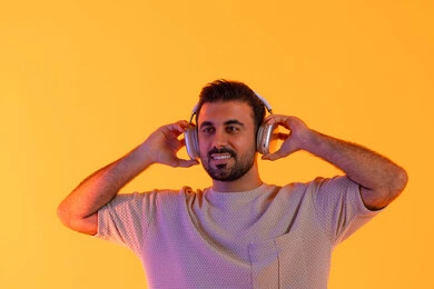 متابعة الموسيقى والترفيه بالهيدفون، الاهتمام بالمظهر الخارجي، تقنية الهيدفون الحديثة، بهدف التسلية  بالاستماع الى الموسيقى العصرية، مفهوم الغناء والطرب، بورتريه مقرب لشاب اسمر عربي خليجي اماراتي مبتسم يرتدي الزي الكاجوال  يضع على رأسه سماعة الرأس، خلفية برتقالي