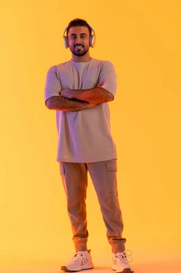 الوقوف بثقة بايدي متقاطعة، الترفيه بسماع المقاطع الصوتية، الاهتمام بالمظهر الخارجي، الاستماع الى الموسيقى العصرية، مفهوم الغناء والطرب، بورتريه مقرب لشاب اسمر عربي خليجي اماراتي مبتسم يرتدي الزي الكاجوال  يضع على رأسه سماعة الرأس، الجسم بالطول الكامل، خلفية برتقالي