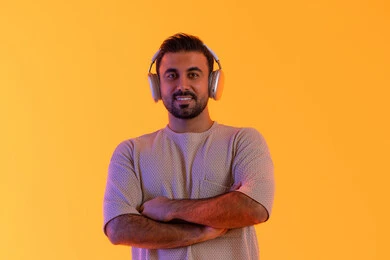 الوقوف بايدي متقاطعة، الاهتمام بالمظهر الخارجي، تقنية الهيدفون الحديثة، بهدف التسلية والمتعة الاستماع الى الموسيقى العصرية، مفهوم الغناء والطرب، بورتريه مقرب لشاب اسمر عربي خليجي اماراتي مبتسم يرتدي الزي الكاجوال  يضع على رأسه سماعة الرأس، خلفية برتقالي