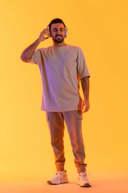 الترفيه بسماع المقاطع الصوتية الهيدفون، الاهتمام بالمظهر الخارجي، الاستماع الى الموسيقى العصرية، مفهوم الغناء والطرب، بورتريه مقرب لشاب اسمر عربي خليجي اماراتي مبتسم يرتدي الزي الكاجوال  يضع على رأسه سماعة الرأس، الجسم بالطول الكامل، خلفية برتقالي