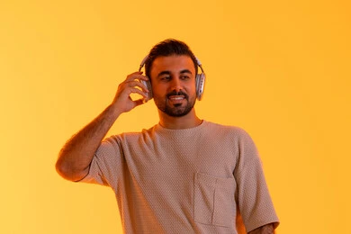 الأناقة و الاهتمام بالمظهر الخارجي، تقنية الهيدفون الحديثة، بهدف التسلية والمتعة الاستماع الى الموسيقى العصرية، مفهوم الغناء والطرب، بورتريه مقرب لشاب اسمر عربي خليجي اماراتي مبتسم يرتدي الزي الكاجوال  يمسك بيده سماعة الرأس بتعابير الفرح والسرور، خلفية برتقالي