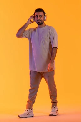 الموسيقى والترفيه بالهيدفون، الاهتمام بالمظهر الخارجي،  بهدف التسلية  بالاستماع الى الموسيقى العصرية، مفهوم الغناء والطرب، بورتريه مقرب لشاب اسمر عربي خليجي اماراتي مبتسم يرتدي الزي الكاجوال  يضع على رأسه سماعة الرأس، الجسم بالطول الكامل، خلفية برتقالي