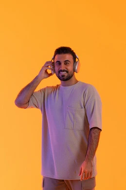 الاهتمام بالمظهر الخارجي، استخدام  تقنية الهيدفون الحديثة، بهدف التسلية والمتعة الاستماع الى الموسيقى العصرية، مفهوم الغناء والطرب، بورتريه مقرب لشاب اسمر عربي خليجي اماراتي مبتسم يرتدي الزي الكاجوال  يمسك بيده سماعة الرأس بتعابير الفرح والسرور، خلفية برتقالي