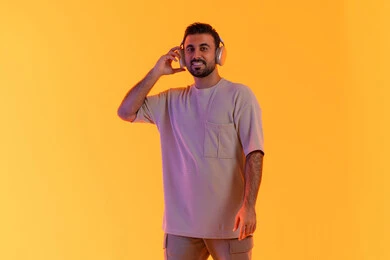 A man using modern headphone technology, for the purpose of entertainment and enjoyment of listening to contemporary music, the concept of singing and melody, a close-up portrait of a smiling young Arab Gulf Emirati man wearing casual attire holding headphones in his hand with expressions of joy and happiness, orange background.