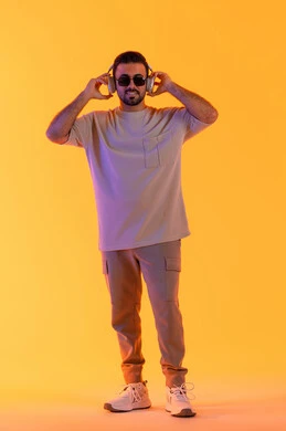 الغناء والطرب الشبابي، قضاء اوقات ممتعة بالاستماع الى الموسيقى، رجل يستخدم تقنية الهيدفون الحديثة، بورتريه لشاب اسمر عربي خليجي اماراتي مبتسم يرتدي الزي الكاجوال والنظارة الشمسية يمسك بيده سماعة الرأس بتعابير الفرح والسرور، صورة الجسم بالكامل، خلفية برتقالي