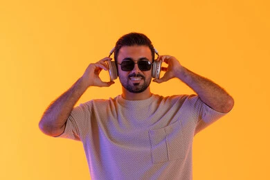 بهدف التسلية والمتعة الاستماع الى الموسيقى العصرية، مفهوم الغناء والطرب، رجل يستخدم تقنية الهيدفون الحديثة، بورتريه مقرب لشاب اسمر عربي خليجي اماراتي مبتسم يرتدي الزي الكاجوال والنظارة الشمسية يمسك بيده سماعة الرأس بتعابير الفرح والسرور، خلفية برتقالي