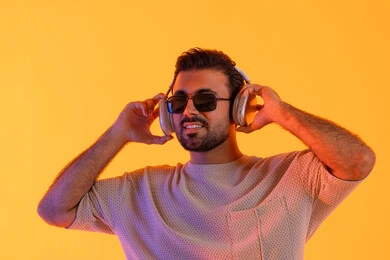 الاستماع الى الموسيقى بهدف التسلية والمتعة، مفهوم الغناء والطرب، رجل يستخدم تقنية الهيدفون الحديثة، بورتريه مقرب لشاب اسمر عربي خليجي اماراتي مبتسم يرتدي الزي الكاجوال والنظارة الشمسية يمسك بيده سماعة الرأس بتعابير الفرح والسرور، خلفية برتقالي