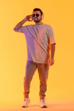 مفهوم الغناء والطرب، قضاء اوقات ممتعة بالاستماع الى الموسيقى، رجل يستخدم تقنية الهيدفون الحديثة، بورتريه لشاب اسمر عربي خليجي اماراتي مبتسم يرتدي الزي الكاجوال والنظارة الشمسية يمسك بيده سماعة الرأس بتعابير الفرح والسرور، صورة الجسم بالكامل، خلفية برتقالي