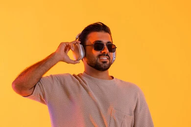 Enjoyable times listening to music, the concept of singing and melody, a man using modern headphone technology, a close-up portrait of a smiling young Arab Gulf Emirati man wearing casual attire and sunglasses, holding a headset in his hand with expressions of joy and happiness, orange background.