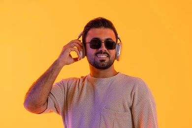 Listening to audio clips, spending enjoyable times listening to music, the concept of singing and melody, a man using modern headphone technology, a portrait of a smiling young Arab Gulf Emirati man wearing casual attire and sunglasses, holding headphones in his hand with expressions of joy and happiness, orange background.