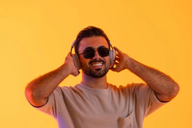 التفاعل والتناغم مع الموسيقى، اوقات ممتعة بالاستماع الى الموسيقى، مفهوم الغناء والطرب، رجل يستخدم تقنية الهيدفون الحديثة، بورتريه لشاب اسمر عربي خليجي اماراتي مبتسم يرتدي الزي الكاجوال والنظارة الشمسية يمسك بيده سماعة الرأس بتعابير الفرح والسرور، خلفية برتقالي