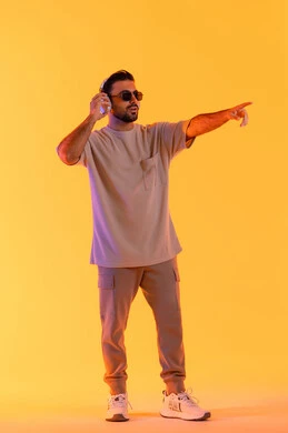 الاشارة لشيء ما، اوقات ممتعة بالاستماع الى الموسيقى، مفهوم الغناء والطرب، رجل يستخدم تقنية الهيدفون الحديثة، بورتريه لشاب اسمر عربي خليجي اماراتي مبتسم يرتدي الزي الكاجوال والنظارة الشمسية يمسك بيده سماعة الرأس بتعابير الفرح والسرور، خلفية برتقالي
