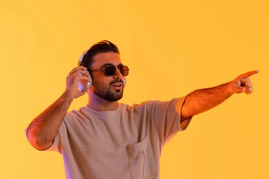 شاب يشير لشيء ما، اوقات ممتعة بالاستماع الى الموسيقى، مفهوم الغناء والطرب، رجل يستخدم تقنية الهيدفون الحديثة، بورتريه لشاب اسمر عربي خليجي اماراتي مبتسم يرتدي الزي الكاجوال والنظارة الشمسية يمسك بيده سماعة الرأس بتعابير الفرح والسرور، خلفية برتقالي