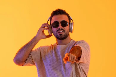 النظر الى الكاميرا والاشارة، اوقات ممتعة بالاستماع الى الموسيقى، مفهوم الغناء والطرب، رجل يستخدم تقنية الهيدفون الحديثة، بورتريه لشاب اسمر عربي خليجي اماراتي مبتسم يرتدي الزي الكاجوال والنظارة الشمسية يمسك بيده سماعة الرأس بتعابير الفرح والسرور، خلفية برتقالي