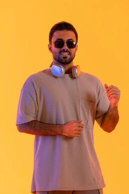 التفاعل والرقص مع الموسيقى، اوقات ممتعة بالاستماع الى الموسيقى، مفهوم الغناء والطرب، رجل يستخدم التقنية الحديثة، بورتريه لشاب اسمر عربي خليجي اماراتي مبتسم يرتدي الزي الكاجوال والنظارة الشمسية يضع سماعة الرأس بتعابير الفرح والسرور، خلفية برتقالي