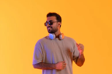 التناغم والتفاعل مع الموسيقى، اوقات ممتعة بالاستماع الى الموسيقى، مفهوم الغناء والطرب، رجل يستخدم التقنية الحديثة، بورتريه لشاب اسمر عربي خليجي اماراتي مبتسم يرتدي الزي الكاجوال والنظارة الشمسية يضع سماعة الرأس بتعابير الفرح والسرور، خلفية برتقالي