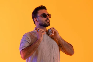 مسك السماعات والنظر بثقة، اوقات ممتعة بالاستماع الى الموسيقى، مفهوم الغناء والطرب، رجل يستخدم التقنية الحديثة، بورتريه لشاب اسمر عربي خليجي اماراتي مبتسم يرتدي الزي الكاجوال والنظارة الشمسية يضع سماعة الرأس بتعابير الفرح والسرور، خلفية برتقالي