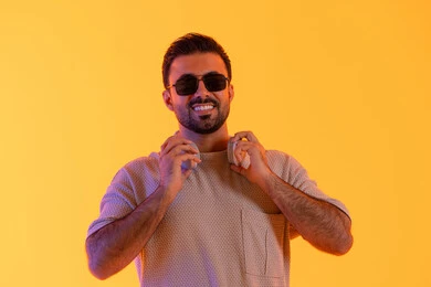 ابتسامة ومسك الهيدفون باليد، اوقات ممتعة بالاستماع الى الموسيقى، مفهوم الغناء والطرب، رجل يستخدم التقنية الحديثة، بورتريه لشاب اسمر عربي خليجي اماراتي مبتسم يرتدي الزي الكاجوال والنظارة الشمسية يضع سماعة الرأس بتعابير الفرح والسرور، خلفية برتقالي