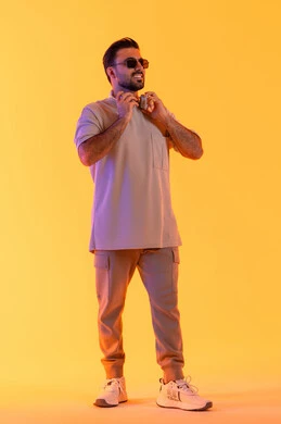 الوقوف بثقة واستقامة ملامح الرجولة، اوقات ممتعة بالاستماع الى الموسيقى، مفهوم الغناء والطرب، رجل يستخدم التقنية الحديثة، بورتريه لشاب اسمر عربي خليجي اماراتي مبتسم يرتدي الزي الكاجوال والنظارة الشمسية يضع سماعة الرأس بتعابير الفرح والسرور، خلفية برتقالي