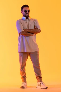Standing with crossed arms and a straight masculine demeanor, enjoying moments listening to music, the concept of singing and melody, a man using modern technology, a portrait of a smiling young Arab Gulf Emirati man with a dark complexion wearing casual attire and sunglasses, wearing headphones with expressions of joy and happiness, orange background.