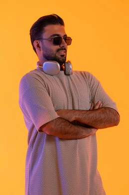 الايدي المتقاطعة واستقامة ملامح الرجولة، اوقات ممتعة بالاستماع الى الموسيقى، مفهوم الغناء والطرب، رجل يستخدم التقنية الحديثة، بورتريه لشاب اسمر عربي خليجي اماراتي مبتسم يرتدي الزي الكاجوال والنظارة الشمسية يضع سماعة الرأس بتعابير الفرح والسرور، خلفية برتقالي