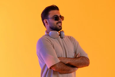 ملامح الرجولة الايدي المتقاطعة واستقامة، اوقات ممتعة بالاستماع الى الموسيقى، مفهوم الغناء والطرب، رجل يستخدم التقنية الحديثة، بورتريه لشاب اسمر عربي خليجي اماراتي مبتسم يرتدي الزي الكاجوال والنظارة الشمسية يضع سماعة الرأس بتعابير الفرح والسرور، خلفية برتقالي