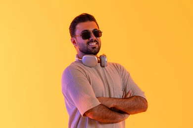 ارتداء سماعات الرأس الصوتية، اوقات ممتعة بالاستماع الى الموسيقى، مفهوم الغناء والطرب، رجل يستخدم التقنية الحديثة، بورتريه لشاب اسمر عربي خليجي اماراتي مبتسم يرتدي الزي الكاجوال والنظارة الشمسية يضع سماعة الرأس بتعابير الفرح والسرور، خلفية برتقالي
