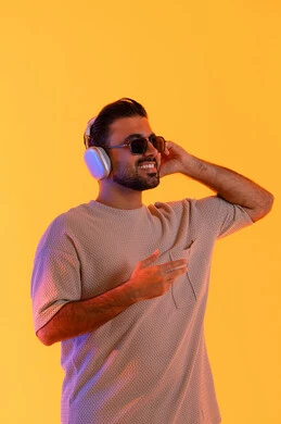 مكالمة هاتفية من خلال سماعات الرأس، اوقات ممتعة بالاستماع الى الموسيقى، مفهوم الغناء والطرب، رجل يستخدم التقنية الحديثة، بورتريه لشاب اسمر عربي خليجي اماراتي مبتسم يرتدي الزي الكاجوال والنظارة الشمسية يضع سماعة الرأس بتعابير الفرح والسرور، خلفية برتقالي