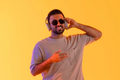 التواصل والتحدث من خلال سماعات الرأس، اوقات ممتعة بالاستماع الى الموسيقى، مفهوم الغناء والطرب، رجل يستخدم التقنية الحديثة، بورتريه لشاب اسمر عربي خليجي اماراتي مبتسم يرتدي الزي الكاجوال والنظارة الشمسية يضع سماعة الرأس بتعابير الفرح والسرور، خلفية برتقالي