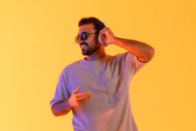 التحدث والتواصل من خلال سماعات الرأس، اوقات ممتعة بالاستماع الى الموسيقى، مفهوم الغناء والطرب، رجل يستخدم التقنية الحديثة، بورتريه لشاب اسمر عربي خليجي اماراتي مبتسم يرتدي الزي الكاجوال والنظارة الشمسية يضع سماعة الرأس بتعابير الفرح والسرور، خلفية برتقالي