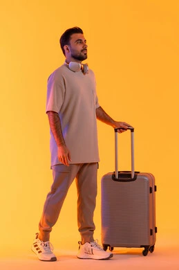Strolling with recreational tours, looking at the camera with gestures of joy and happiness, preparing for an enjoyable summer trip, the concept of tourism during the summer vacation, a portrait of an Arab Gulf Emirati man wearing casual attire and headphones holding a luggage bag in his hand, a full-body image, with an orange background.