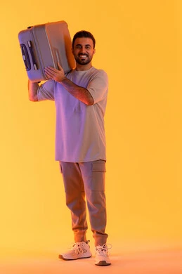 Preparing for an enjoyable summer trip, engaging in recreational tours, looking with gestures of joy and happiness, the concept of tourism during the summer vacation, a portrait of an Arab Gulf Emirati man wearing casual attire carrying a luggage bag on his shoulder, tourism during the summer vacation, orange background.
