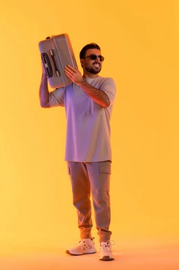 إيماءات السرور والسعادة، التحضير قضاء رحلة صيفية ممتعة، القيام بالجولات الترفيهية، مفهوم السياحة في إجازة الصيف، بورتريه لرجل عربي خليجي اماراتي يرتدي الزي الكاجوال ونظارة شمسية يحمل على كتفه حقيبة الامتعة، السياحة في إجازة الصيف, خلفية برتقاليه