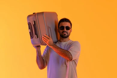 Spending enjoyable times during the vacation, the concept of tourism in the summer vacation, gestures of joy and happiness, preparing for an enjoyable summer trip, engaging in recreational tours, a close-up portrait of an Emirati Gulf Arab man wearing casual attire and sunglasses, carrying a luggage bag on his shoulder, with an orange background.